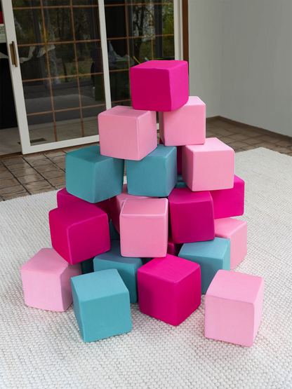 A pyramid-like stack of soft cubes in bright pink, light pink, and teal sits on a white textured rug indoors, near glass doors with a view outside.